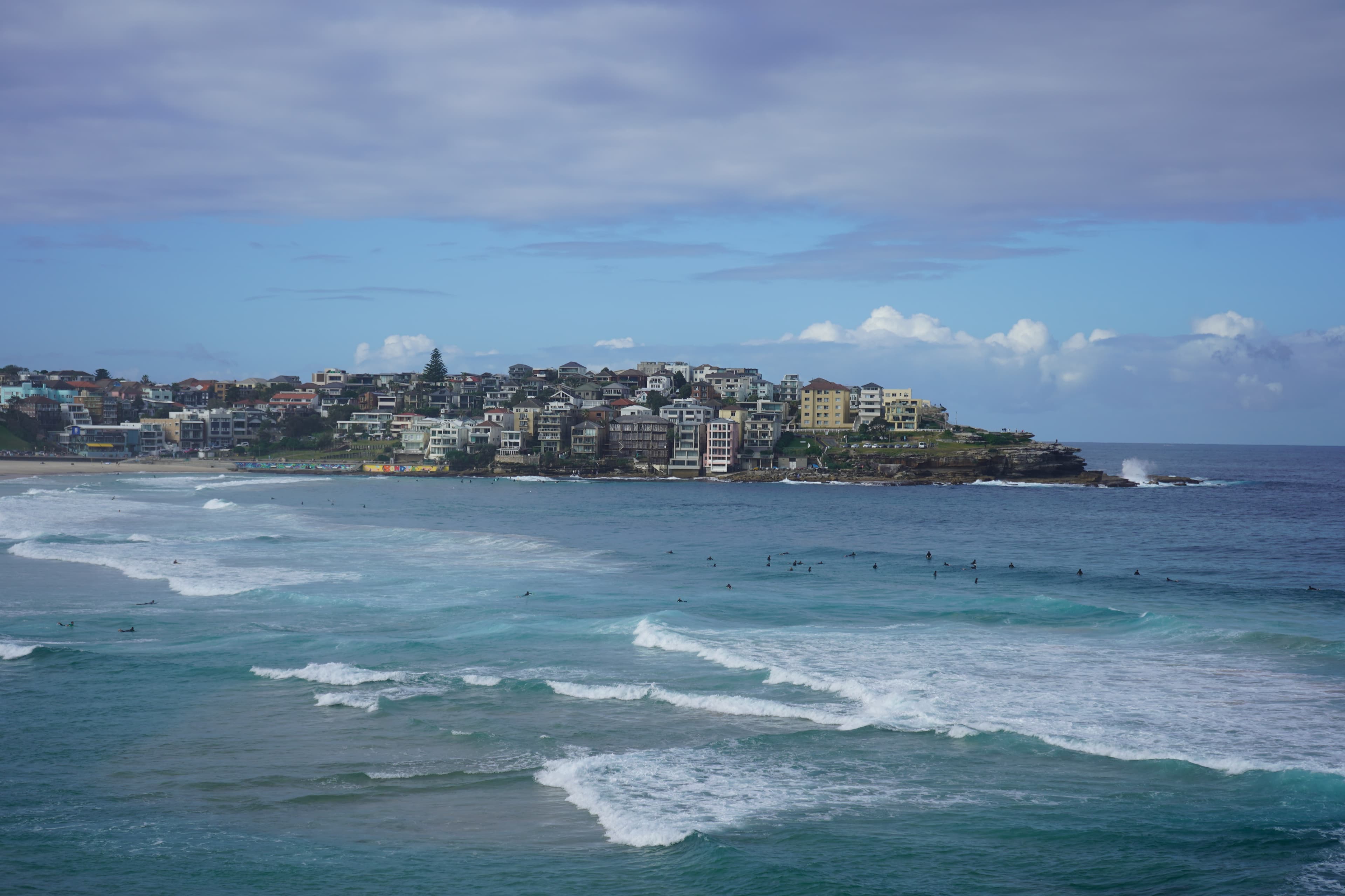 Waves and surfers below a coastal neighbourhood under a cloudy blue sky