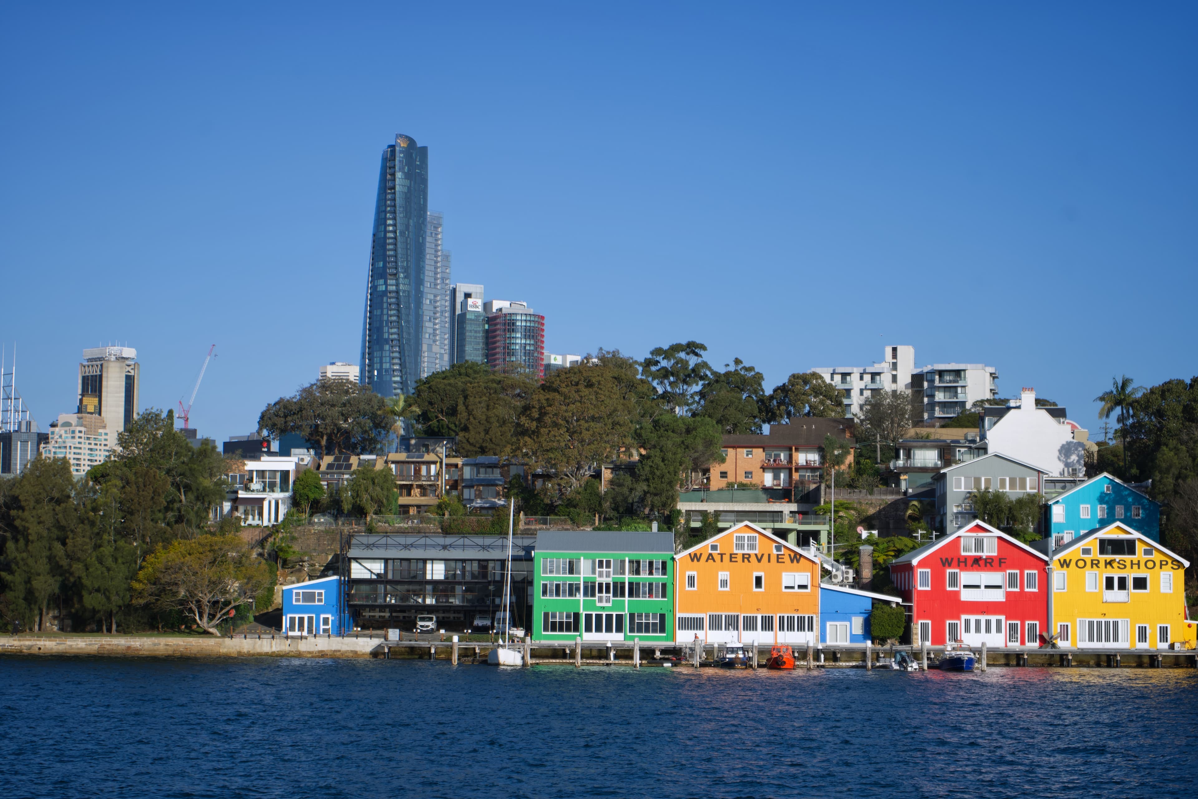 Colourful waterfront buildings beneath trees and a tall city tower across blue harbour water