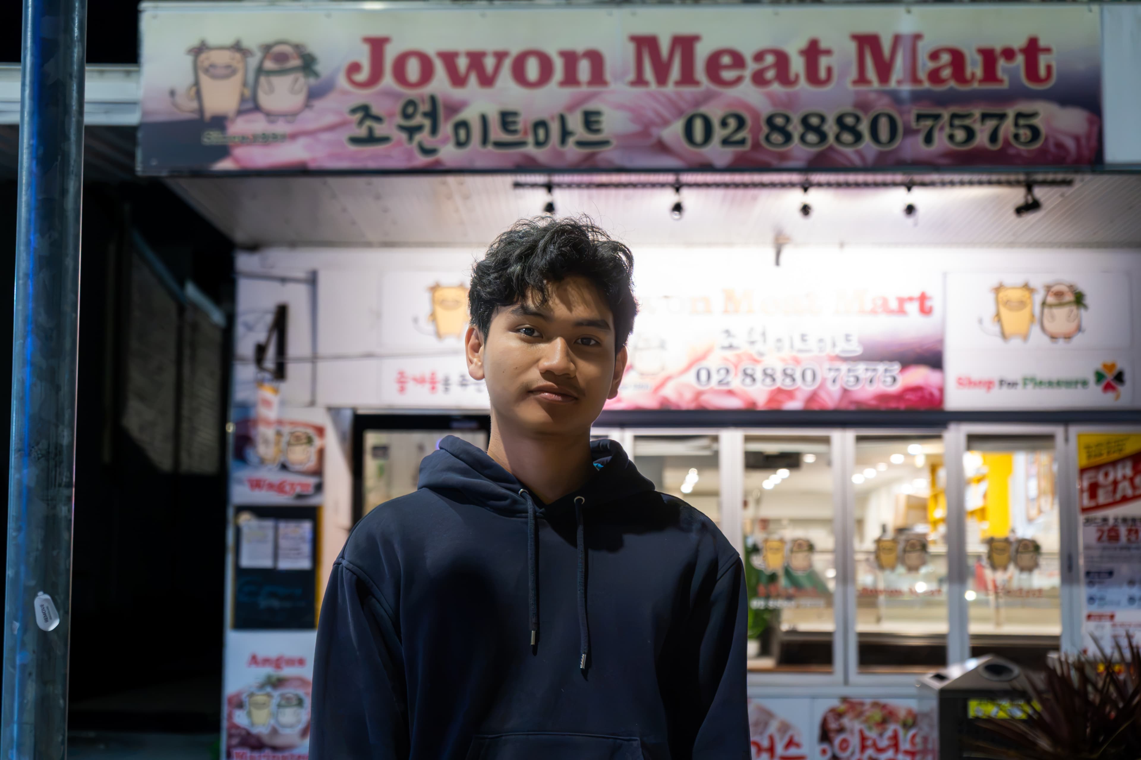 Portrait of a person in a dark hoodie outside an illuminated shopfront at night