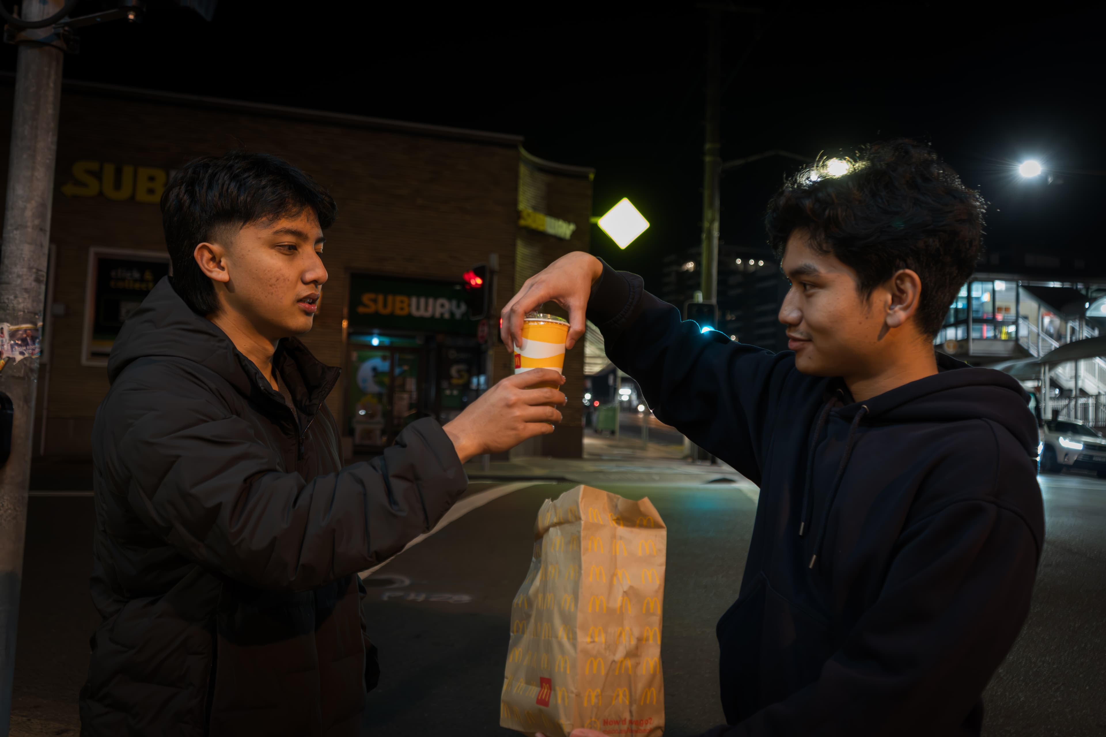 Two people holding a takeaway drink on a street lit by shopfronts at night